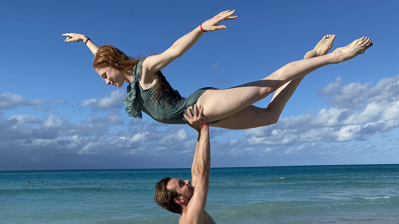 A man lifts a woman above him in a dancers pose with crystal blue waters behind them