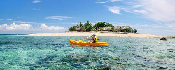 A person in a yellow kayak on sparkly blue, clear waters. A small tropical island with a beach and palm trees is in the background. 