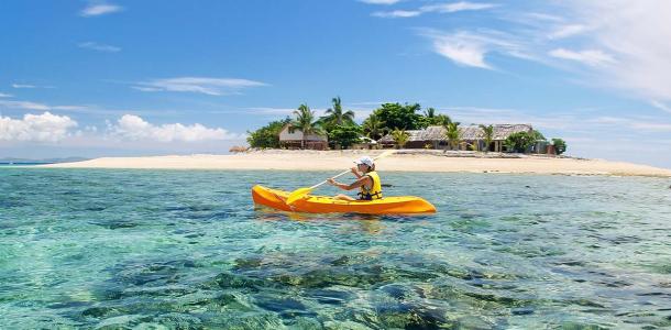 A person in a yellow kayak on sparkly blue, clear waters. A small tropical island with a beach and palm trees is in the background. 